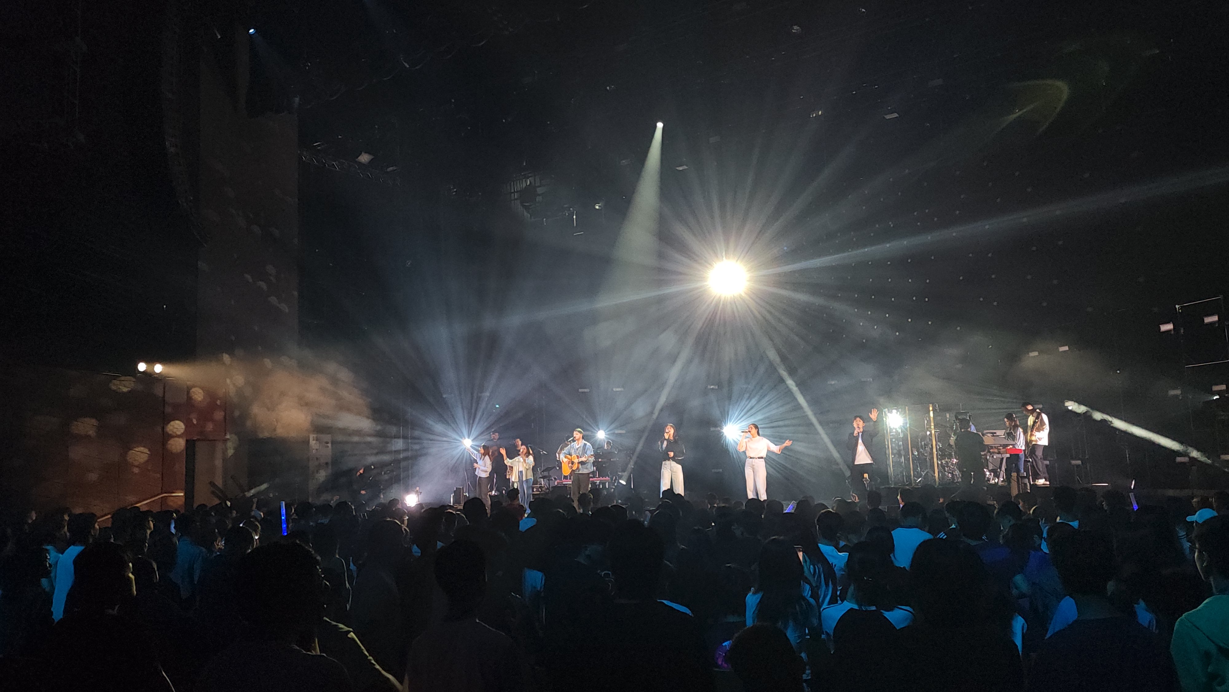A wide concert stage framed by beams of white light and the crowd